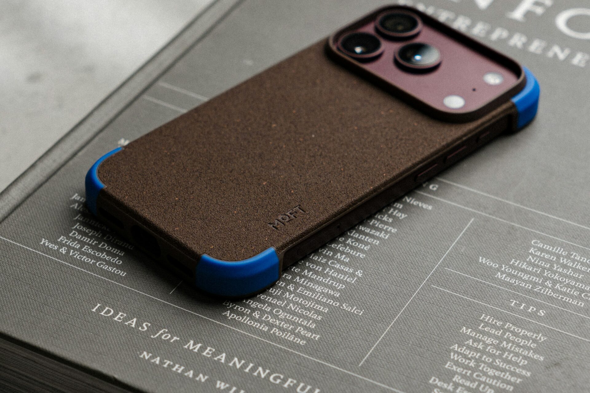 A brown textured phone case with blue corners resting on a grey hardcover book